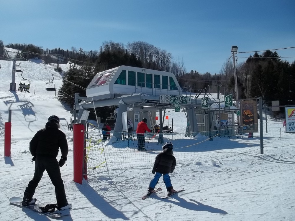 Un planchiste et un skieur se dirigeant vers les télésièges avec leur équipement de location au Mont Lac-Vert à Hébertville.